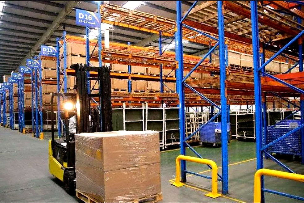 A forklift moving a stretch-wrapped pallet down an aisle of blue and orange pallet racking.